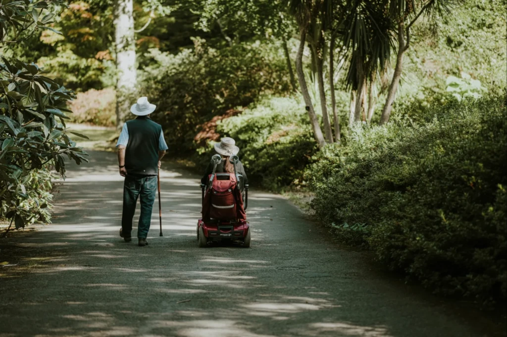 Elderly couple with walking stick and wheelchair going through park